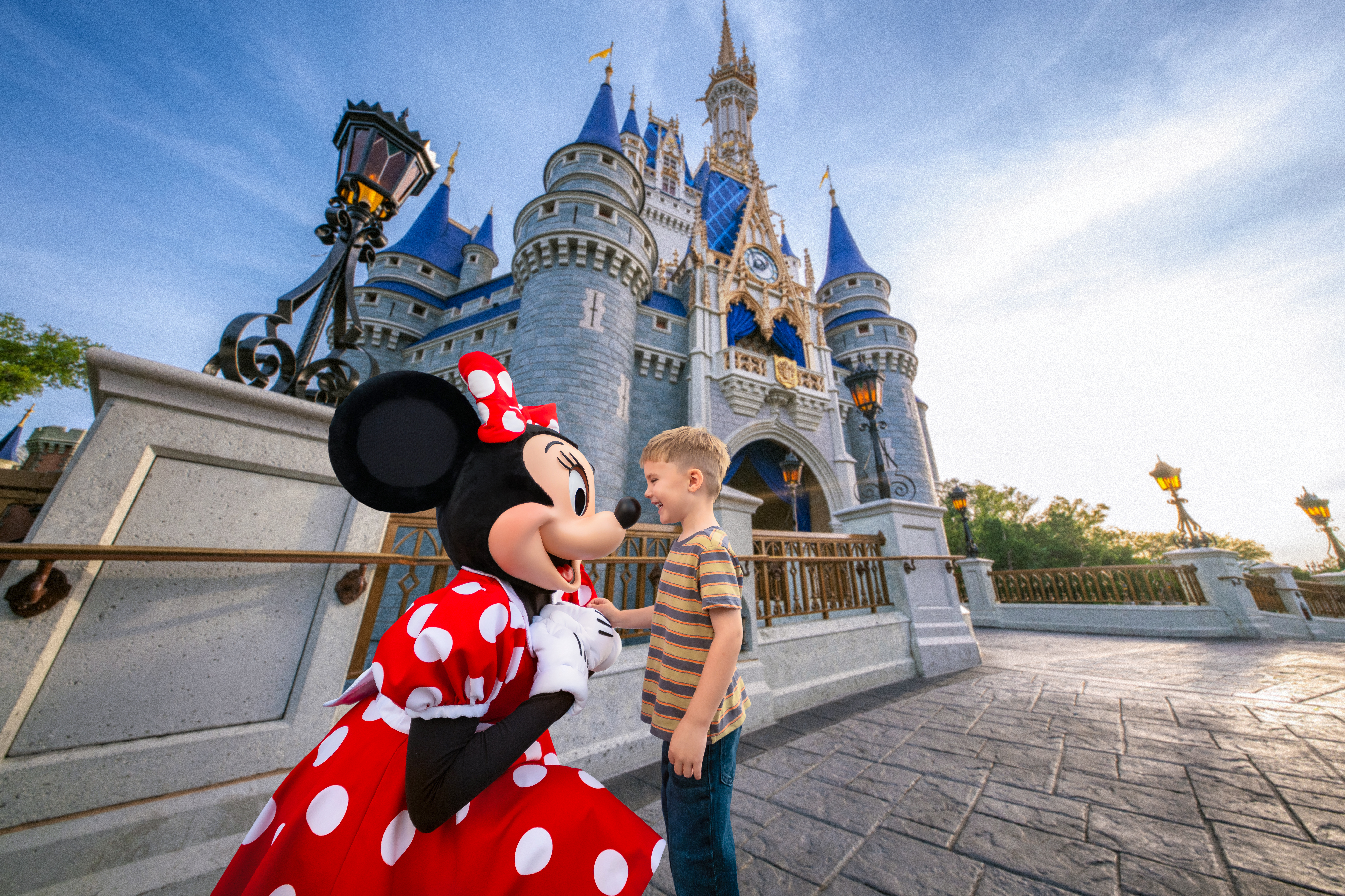 Minnie Mouse is kneeling down to eye level with a little boy in front of Cinderella Castle